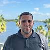 A man stands by a waterfront with palm trees in the background.