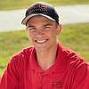 A young man in a red shirt and baseball cap smiles warmly at the camera.