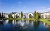 A serene view of an apartment complex with a pond and fountain under a clear blue sky.