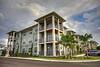 Modern apartment building with balconies and palm trees under a cloudy sky.